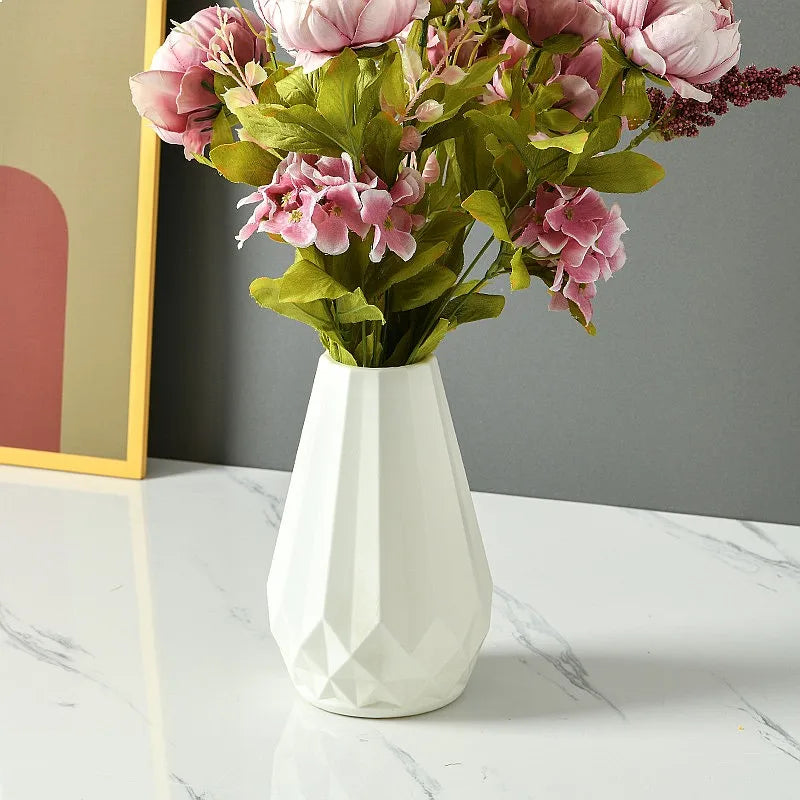A white faceted ceramic vase filled with pink artificial flowers and green leaves, placed on a white marble surface against a gray wall.