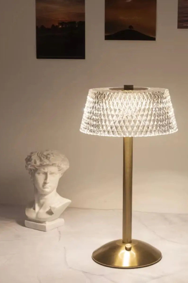 A table lamp with a textured, faceted clear shade and a brushed brass gold base, illuminated on a marble tabletop with decorative props including a classical bust and a vase of flowers.