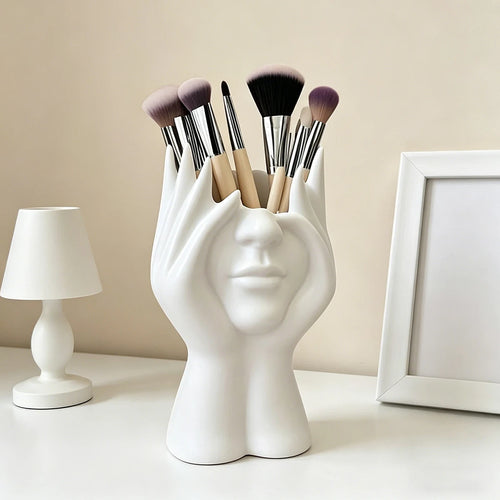 White face-shaped brush holder with makeup brushes on a neutral background