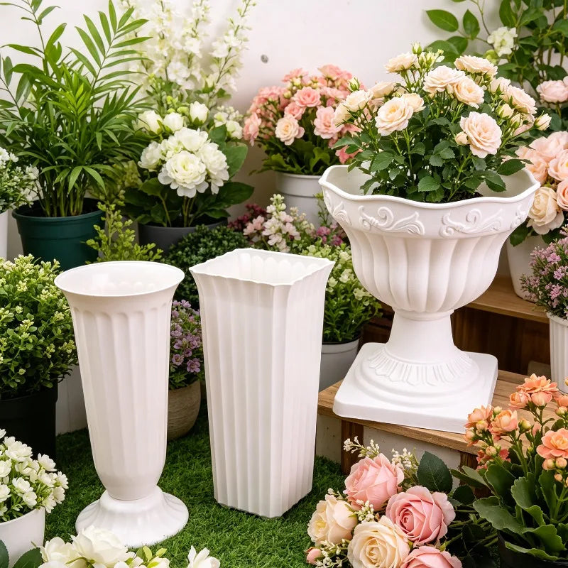 Three white decorative plastic plant pots displayed on artificial grass with a background of potted plants and flowers. The set includes a tall fluted pedestal vase, a tall rectangular planter, and a wide fluted urn pot filled with faux roses.