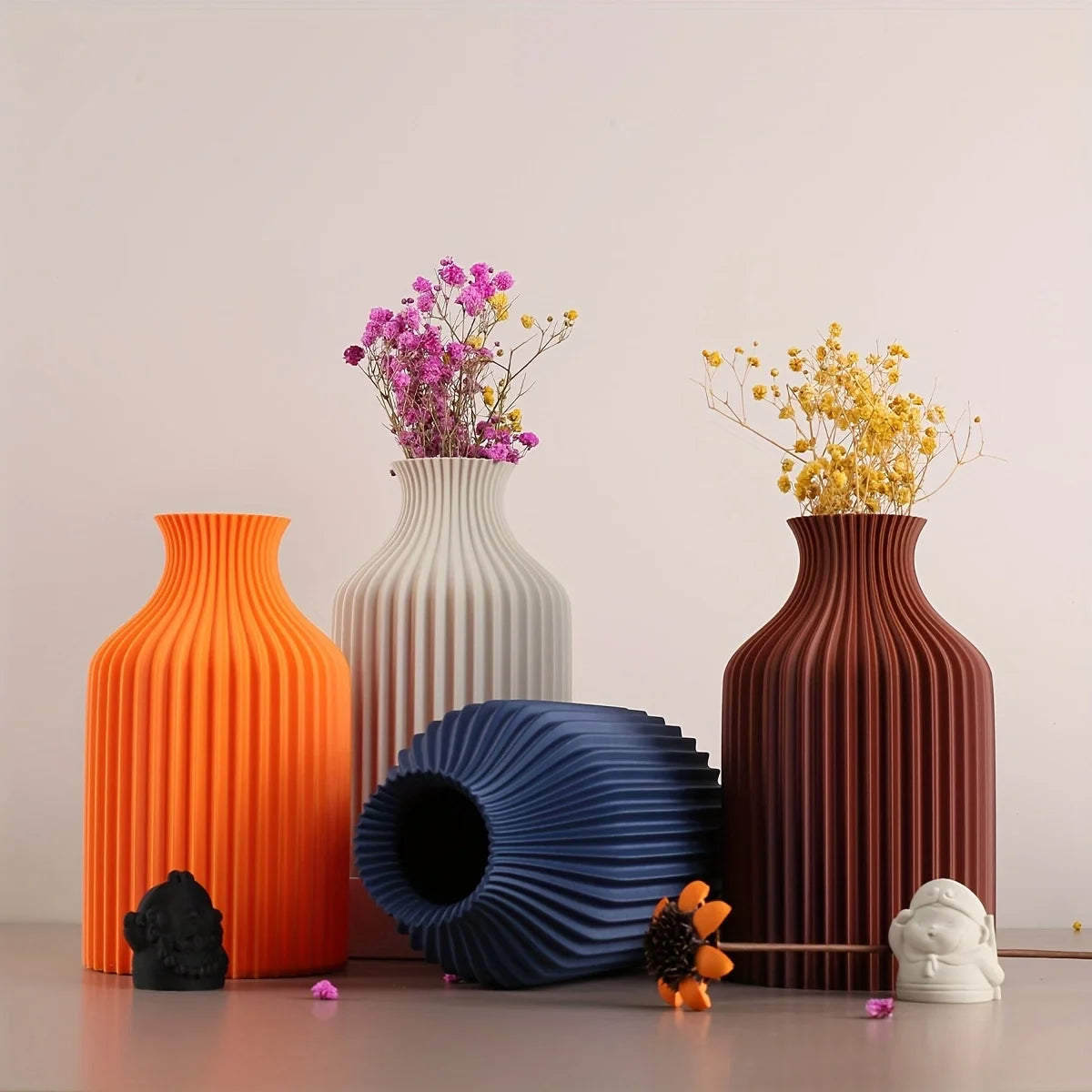 Three ribbed ceramic vases in orange, cream, and reddish-brown, with dried pink and yellow flowers, alongside a blue ribbed vase and small decorative figurines, arranged on a tabletop against a plain light background.