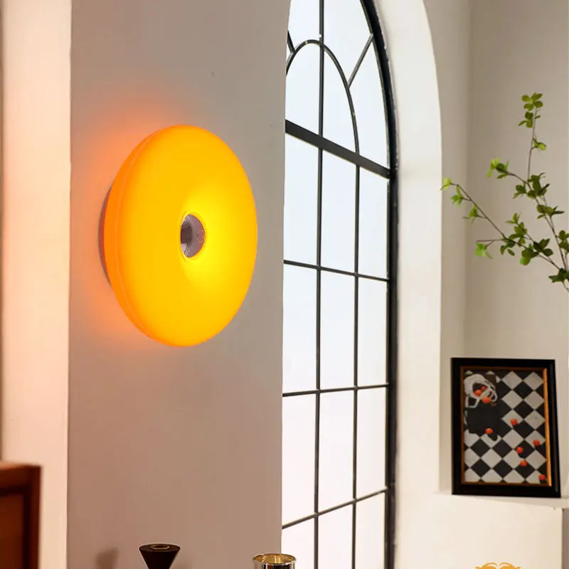 A round, disc-shaped wall sconce light fixture glowing with a warm yellow-orange light, mounted on a white wall next to an arched window.