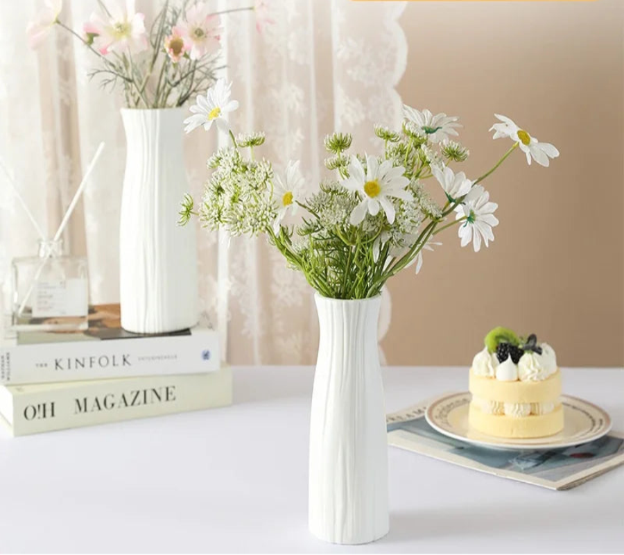 White vase with flowers on a table with books and a small cake.