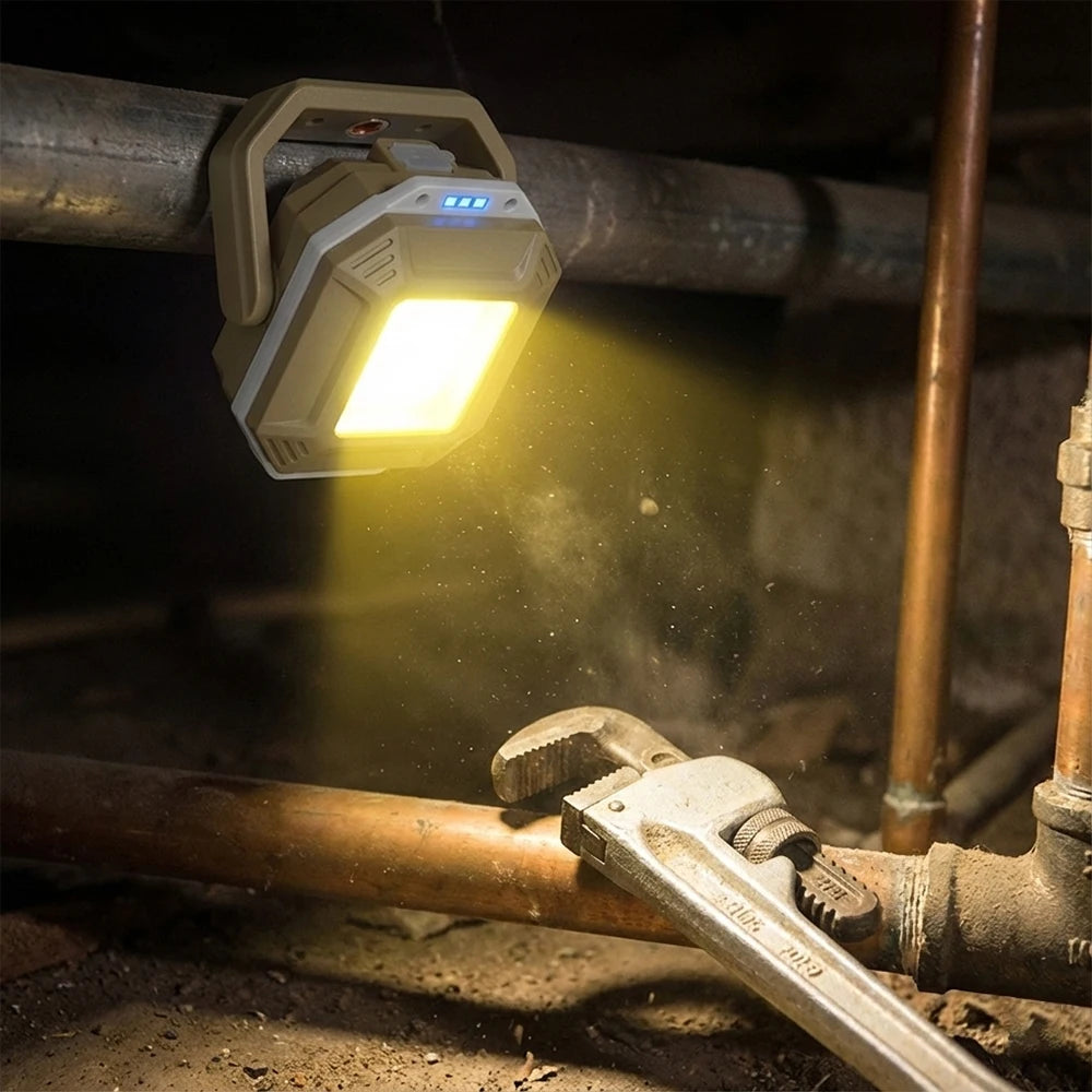 A rugged waterproof work light with a bright yellow glow, mounted on a metal pipe in a dark utility setting with copper pipes and a wrench in the foreground.