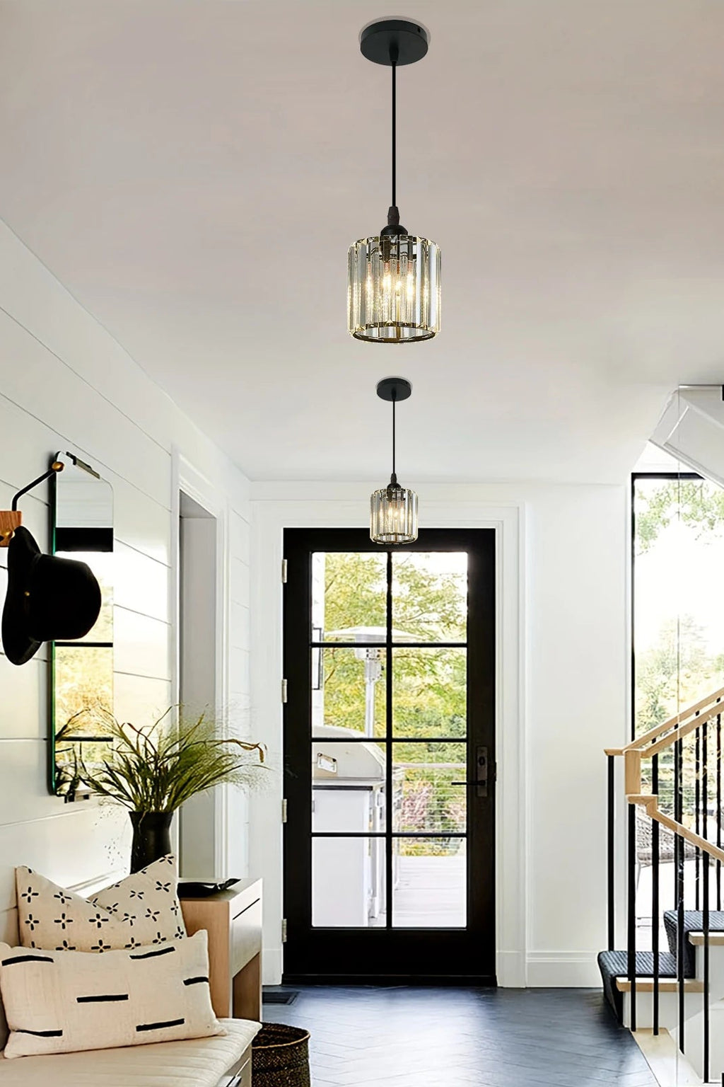 Two modern pendant lights with clear ribbed glass cylindrical shades and black metal hardware hang over a hallway leading to a black-framed glass door. The scene includes a staircase, wood cabinetry, and a bench with pillows in a bright, contemporary interior.