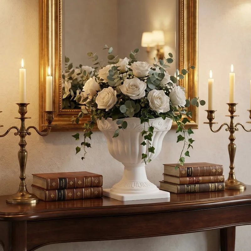 A white classical-style urn vase filled with white roses and green eucalyptus stems, displayed on a dark wooden console table. The setting is decorated with two brass candlesticks, a stack of vintage books on each side, and a large gold-framed mirror in the background.