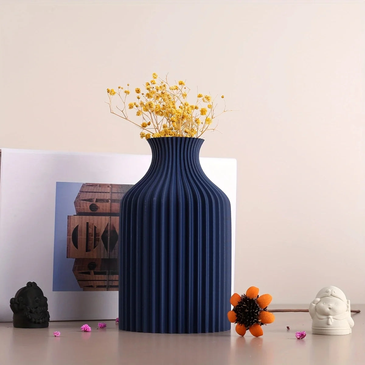 A ribbed, pleated ceramic vase in deep navy blue, filled with yellow baby's breath flowers, styled on a tabletop with a picture frame, small decorative objects, and a white flower in the background.