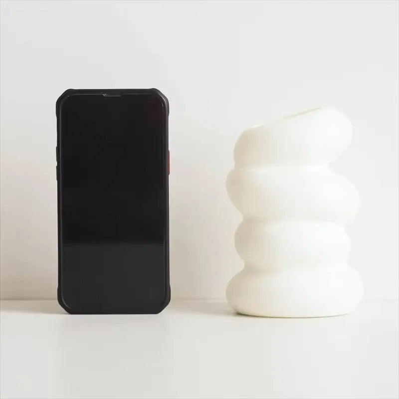 Black smartphone next to a white ceramic vase on a light surface with a white background