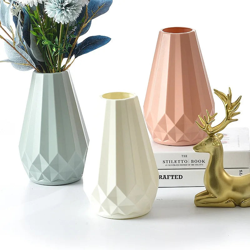 Three modern geometric ceramic vases in pale blue, cream, and blush pink, styled with dried flowers, books, and a gold deer figurine on a white surface.