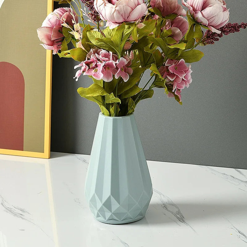 A faceted light blue ceramic vase filled with pink artificial flowers and green leaves, placed on a white marble surface against a gray wall.