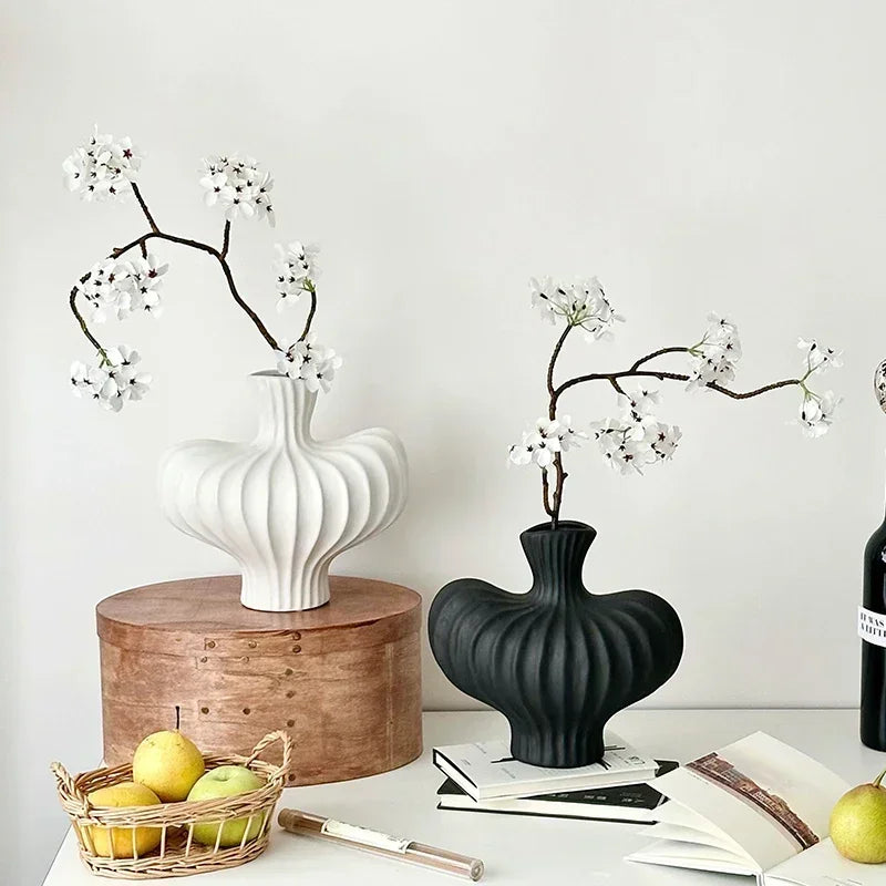 Two ribbed ceramic flower vases, one white and one black, holding white blossoms, styled on a white table with a wooden cylinder, books, and fruit against a light wall.