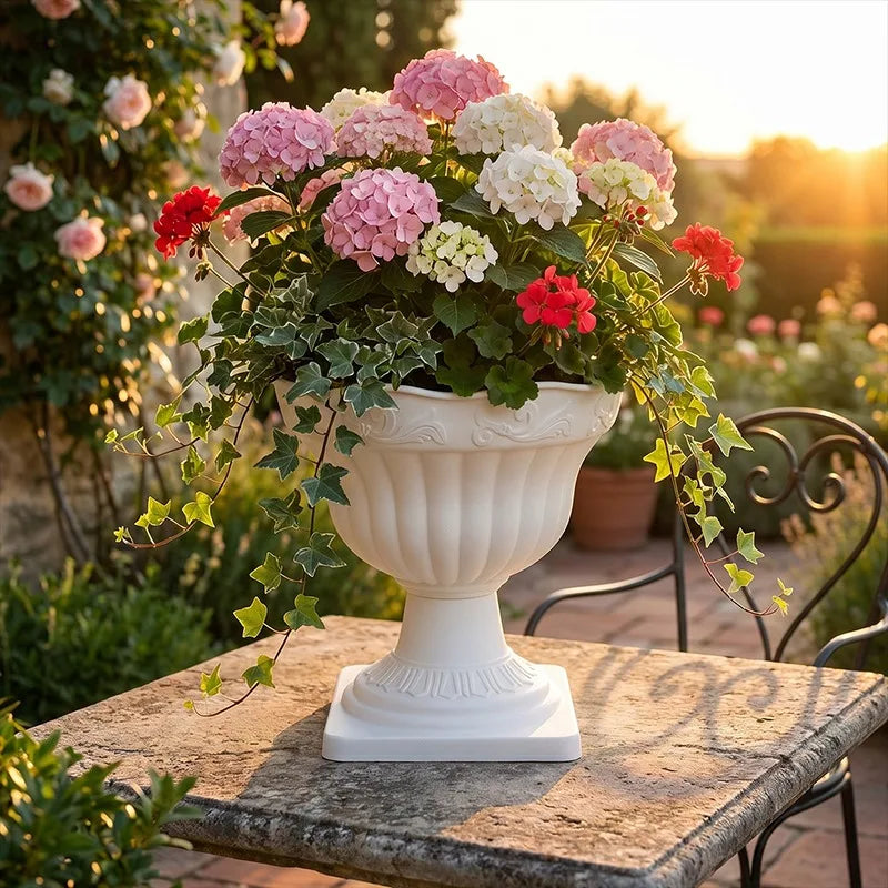 A white classical-style urn planter filled with pink, white, and red hydrangeas and trailing ivy, displayed on a stone table in a sunlit garden.
