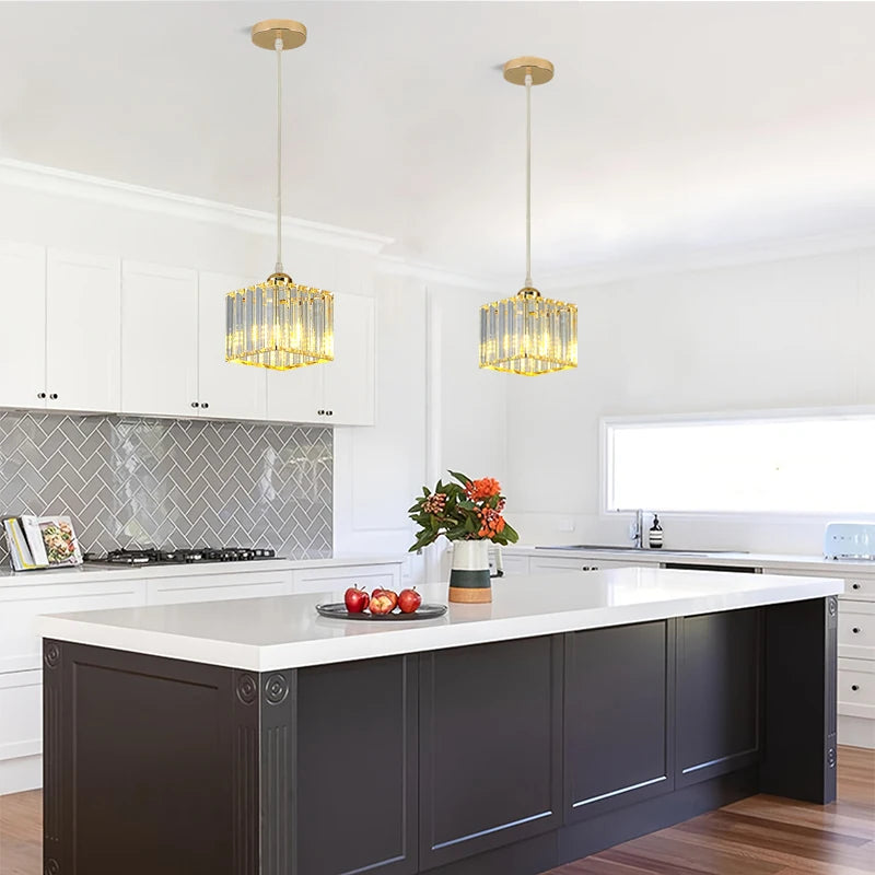 Two modern cylindrical pendant lights with gold-toned frames and clear glass or acrylic vertical bars, hanging over a white kitchen island in a bright, contemporary kitchen.