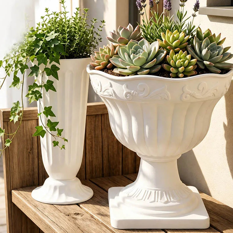 Two white decorative ceramic planters displayed on a wooden shelf. The larger planter on the right is footed and filled with succulents, while the smaller planter on the left is fluted and holds trailing green ivy.