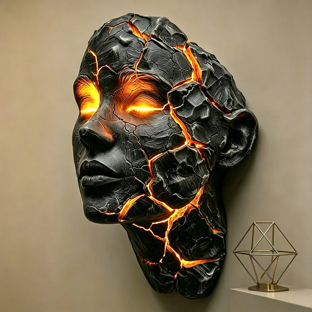 A sculptural black stone-like head with glowing orange fissures and bright orange eyes, mounted on a beige wall above a small gold geometric table lamp.