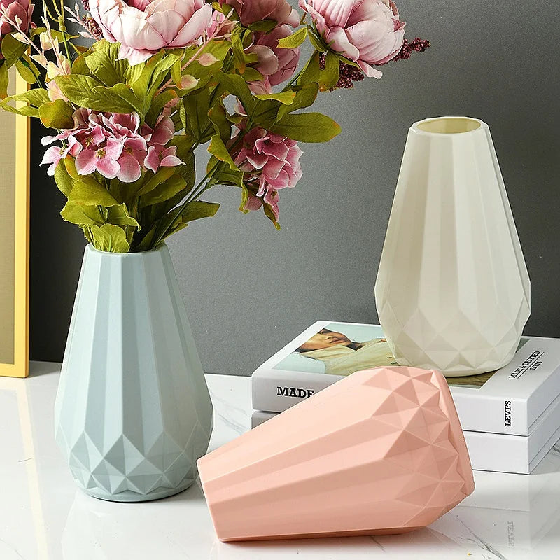 Three faceted geometric ceramic vases in pale blue, blush pink, and cream, styled with artificial pink flowers and books on a white surface against a gray wall.