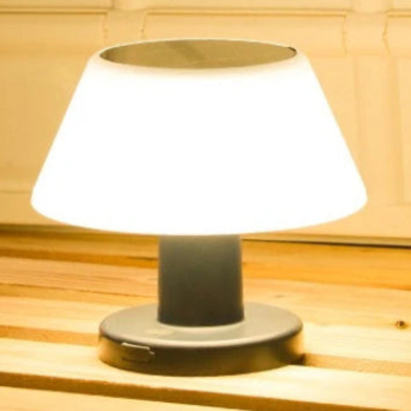 A small table lamp with a glowing white conical shade and a dark gray cylindrical base, shown on a wooden surface against a light wood paneled background.
