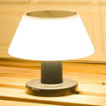 A small table lamp with a glowing white conical shade and a dark gray cylindrical base, shown on a wooden surface against a light wood paneled background.