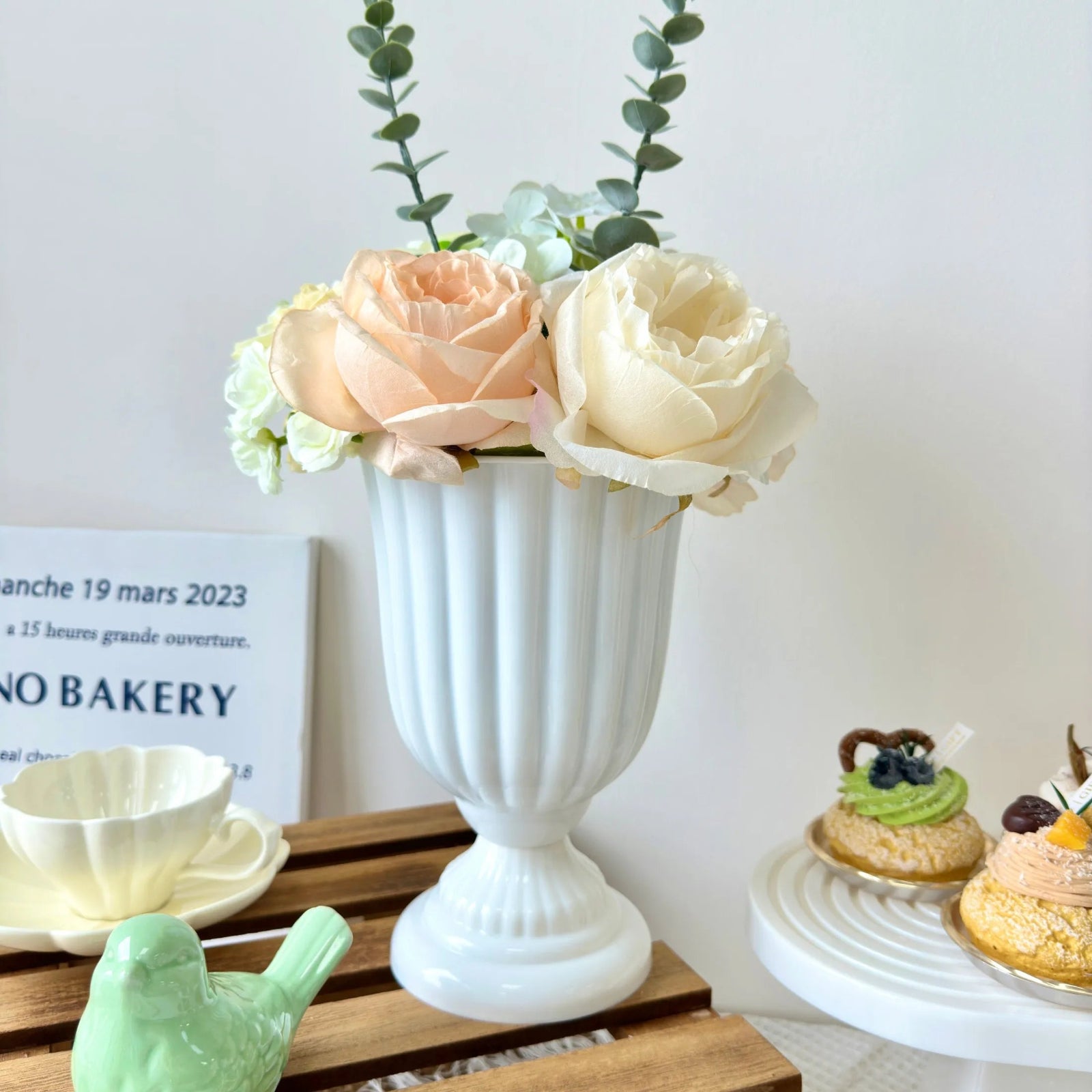 White vase with flowers on a wooden table with pastries in the background