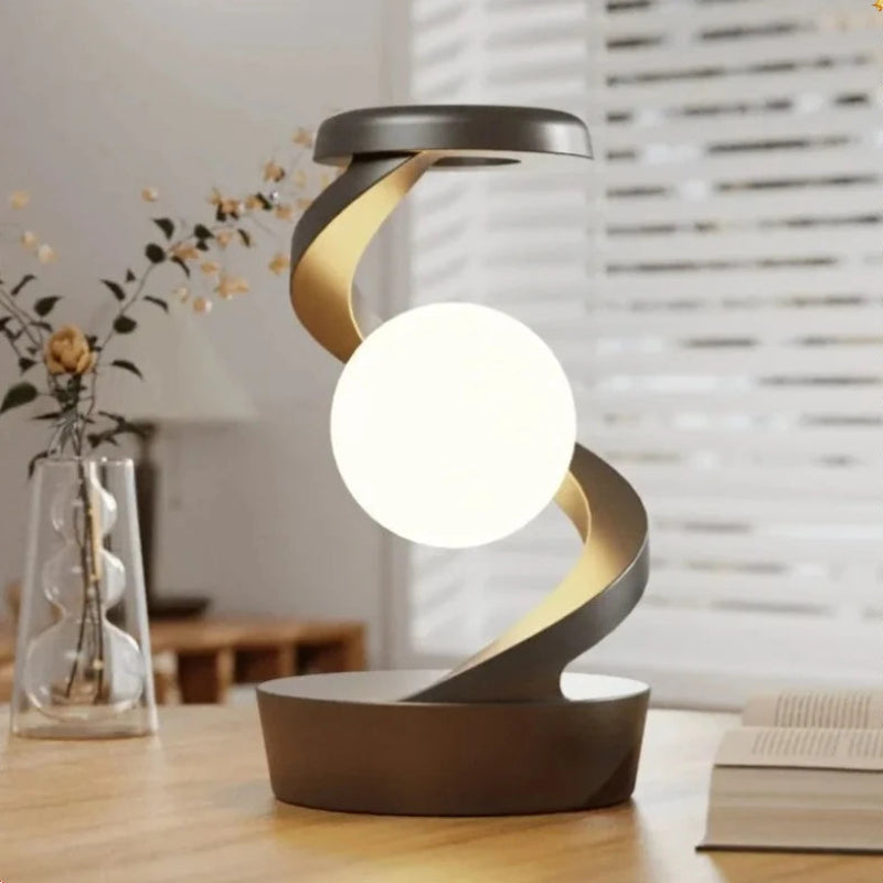 A modern spiral-shaped table lamp with a glowing white oval light, set on a wooden table in a softly lit room with a blurred window and vase in the background.
