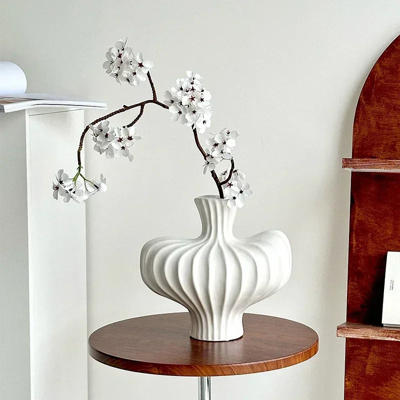 A ribbed white ceramic vase with a curved, sculptural form sits on a round wooden table, holding a flowering branch with white blossoms. The vase is styled in a minimalist interior setting with a white wall, a wooden shelf on the right, and a white shelf on the left.