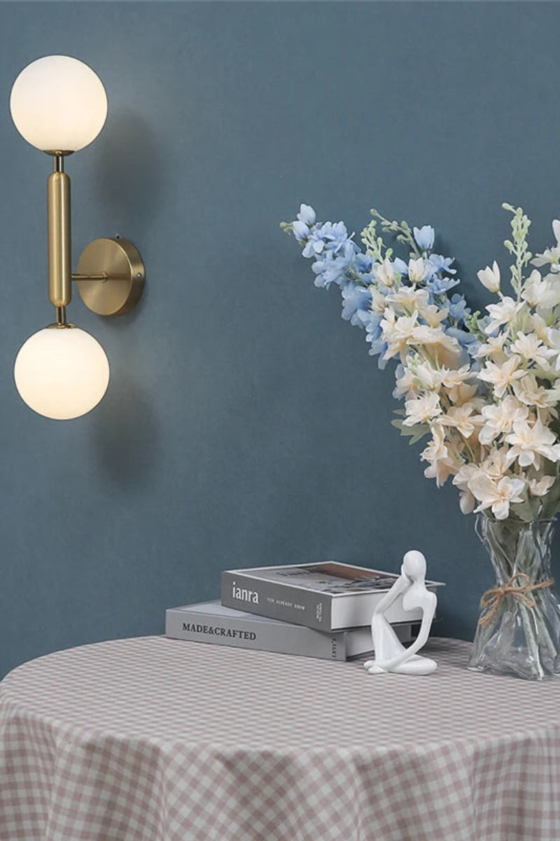 Wall sconce with two round lights on a dark blue wall, next to a table with a checkered tablecloth, books, a small statue, and a vase of flowers.