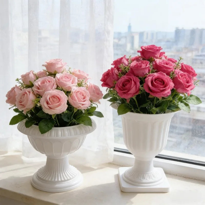 Two white classical-style pedestal flower vases filled with rose bouquets, one in light pink and one in deep pink, placed on a windowsill with sheer curtains and a blurred city view in the background.