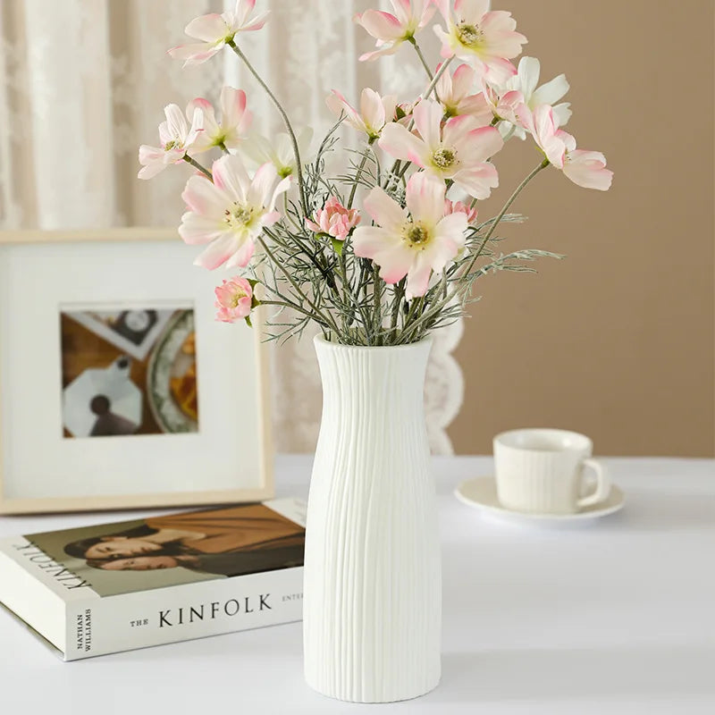 White vase with pink flowers on a table next to books and a cup