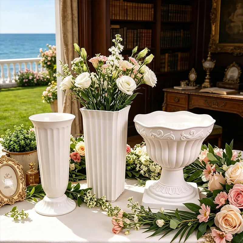 Three white decorative classical-style flower vases arranged on a table in an elegant indoor setting with a view of the ocean outside. The set includes a fluted pedestal vase, a ribbed rectangular vase with a bouquet of white and pink flowers, and a wide scalloped bowl-shaped vase surrounded by additional flowers and greenery.
