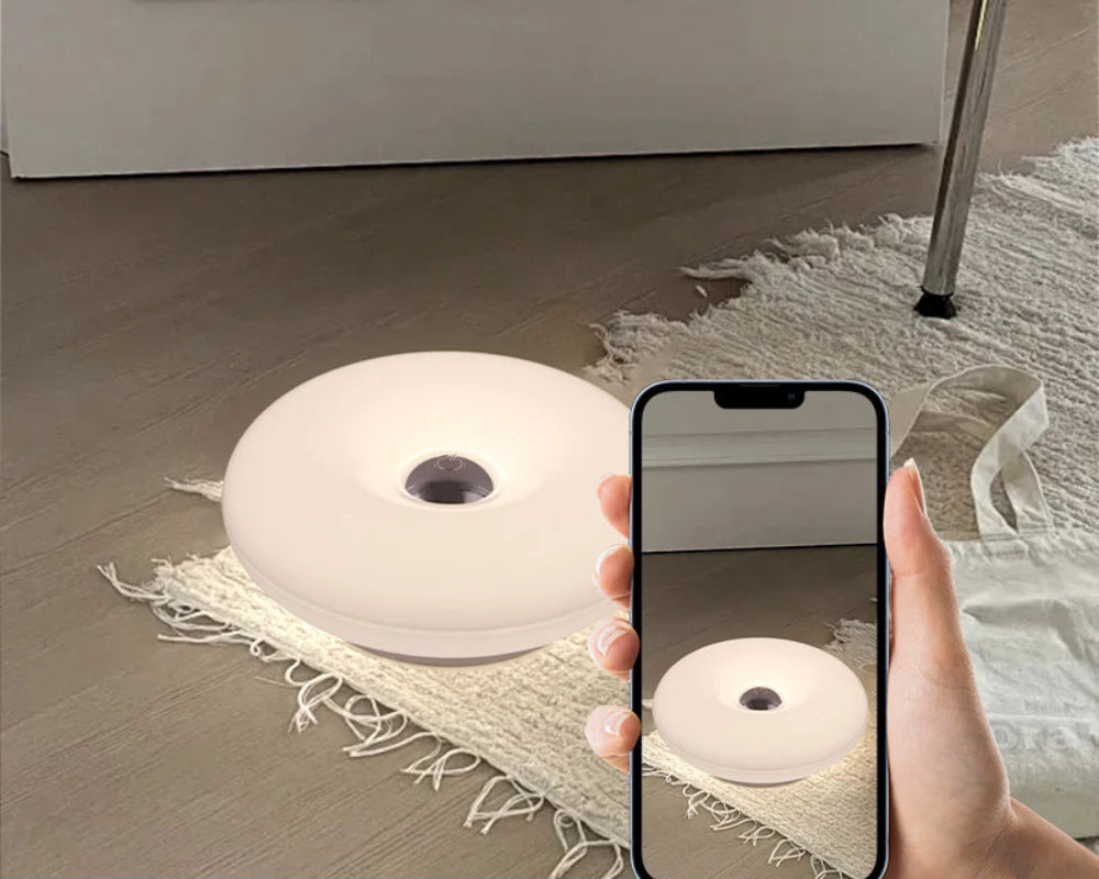 A hand holds a smartphone showing a mirror image of a round, low-profile white light fixture placed on a woven rug over a wood floor. Text at the bottom reads 'No video flashing' and 'The light is soft and the atmosphere is pleasant.