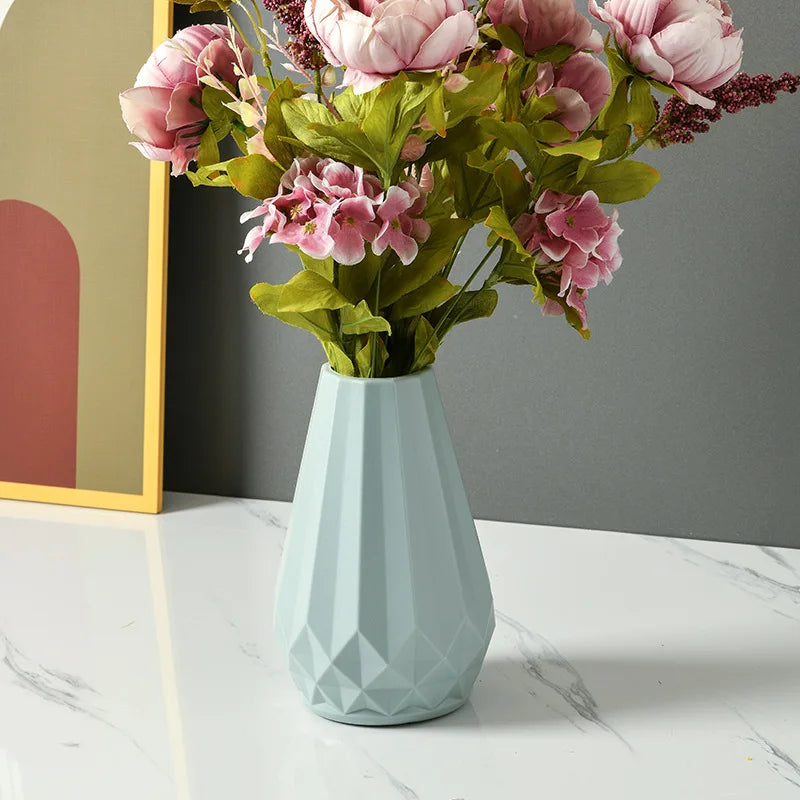 A light blue faceted ceramic vase filled with pink artificial flowers and green leaves, placed on a white marble surface against a gray wall.
