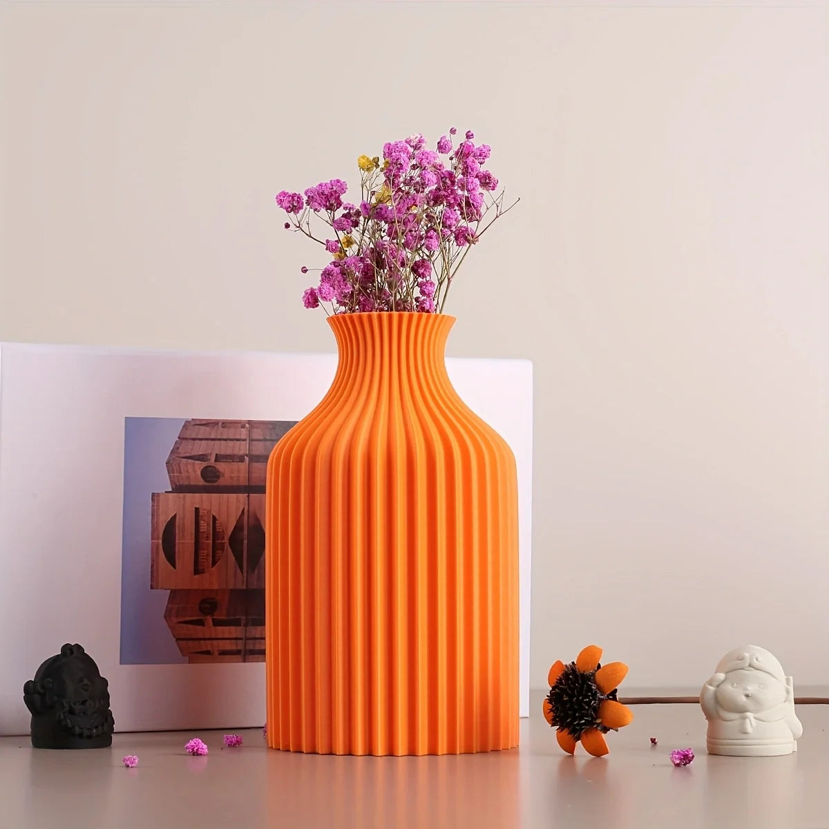 An orange ribbed ceramic vase filled with pink baby's breath flowers, styled on a tabletop with a white picture frame, a small black figurine, a small orange flower, and a white ceramic character against a plain beige wall.