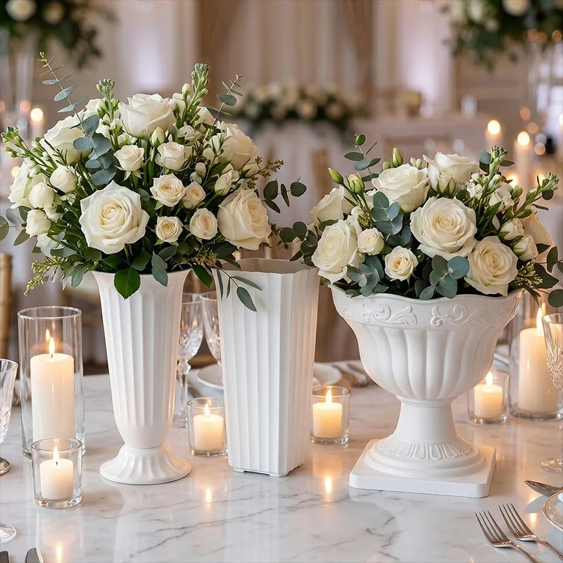 Three white floral centerpieces with white roses, green foliage, and eucalyptus stems displayed in white pedestal vases on a marble table with lit candles.