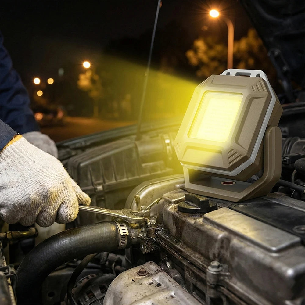 A portable work light with a rugged beige and gray housing is mounted on an open car engine bay at night, casting a bright yellow beam of light onto the engine. A person wearing a blue jacket and white work gloves is visible on the left, using a wrench.