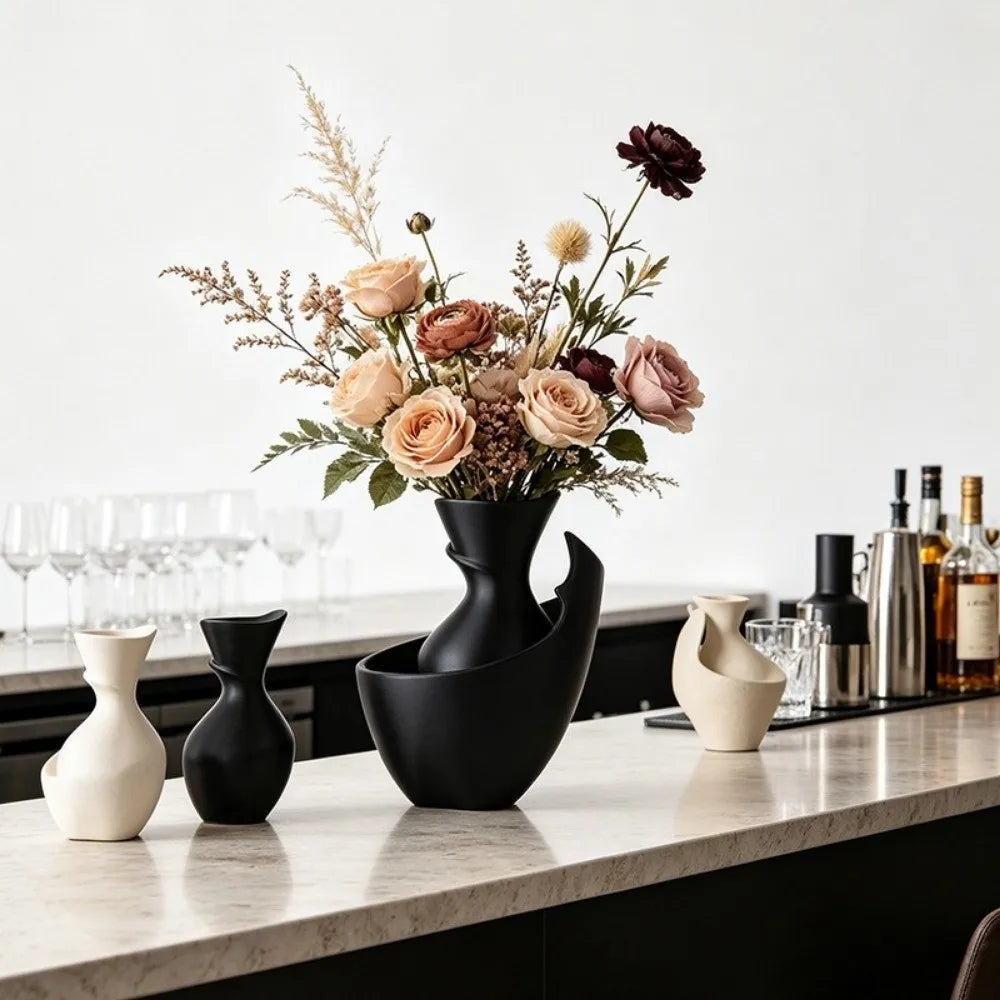 A modern sculptural black ceramic vase filled with a dried floral arrangement, styled on a marble countertop alongside other minimalist vases in a bright kitchen or bar setting.