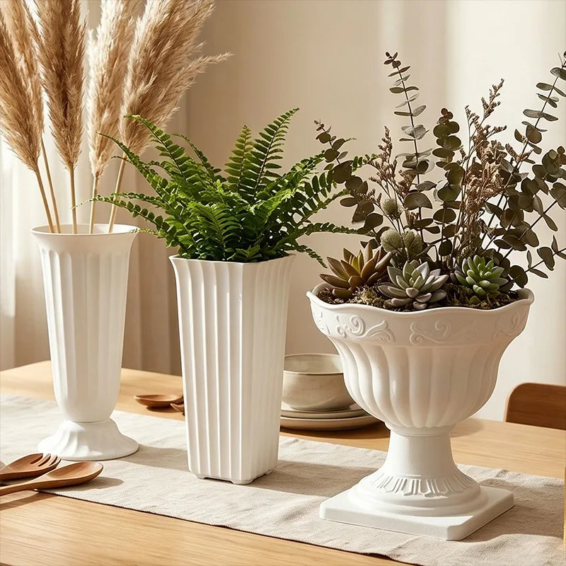 Three white ceramic planters arranged on a wooden table in a warm, styled interior setting. The left planter holds dried pampas grass, the middle planter holds a green fern, and the right planter holds a succulent and eucalyptus arrangement.