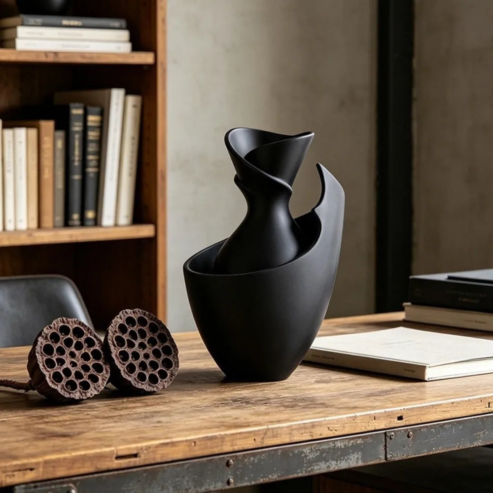 A matte black sculptural vase with a twisted neck and open curved sides, displayed on a rustic wooden table in a styled interior setting with books and lotus pods.