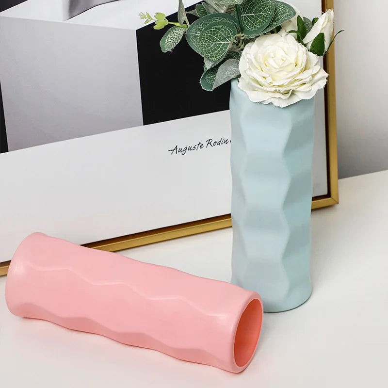 Two ceramic vases, one pink and one light blue, on a surface with a framed picture in the background.