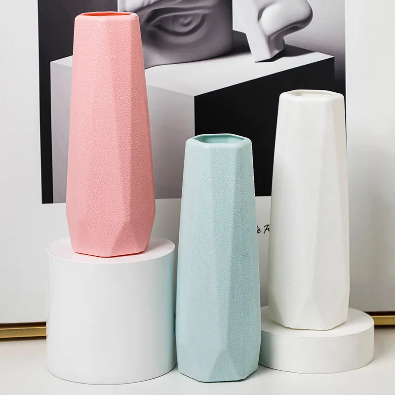 Three geometrically shaped vases in pink, green, and white on a white surface with a black and white abstract art piece in the background.