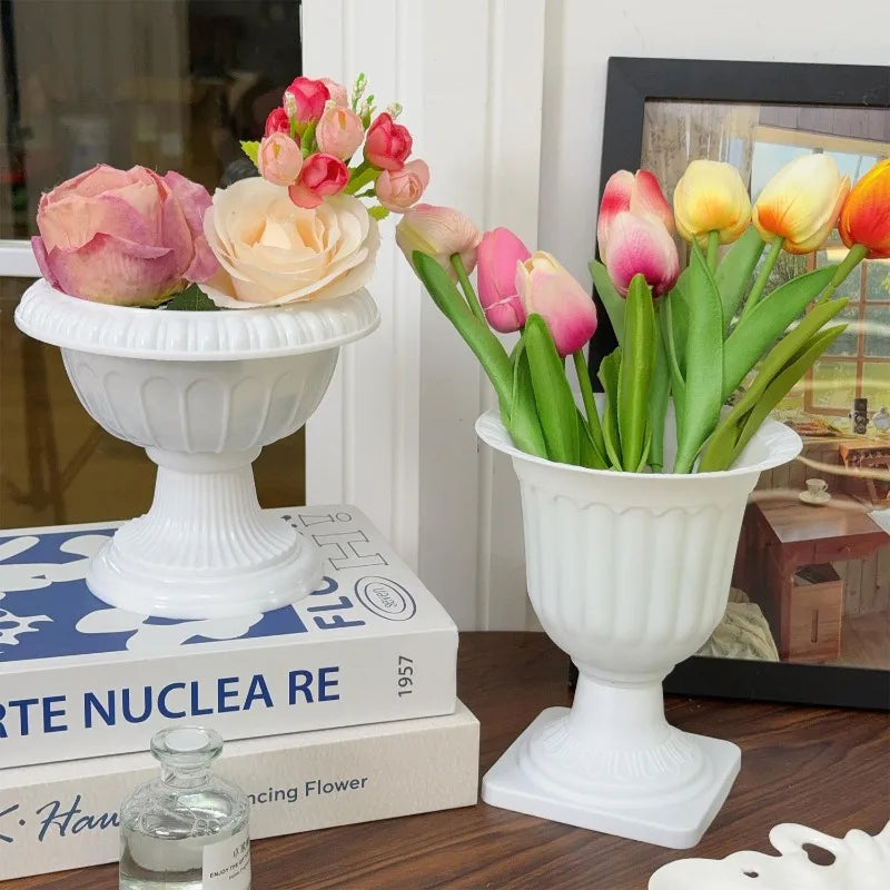 Two white classical-style pedestal flower vases filled with artificial flowers, displayed on a wooden table with books and a picture frame in the background.