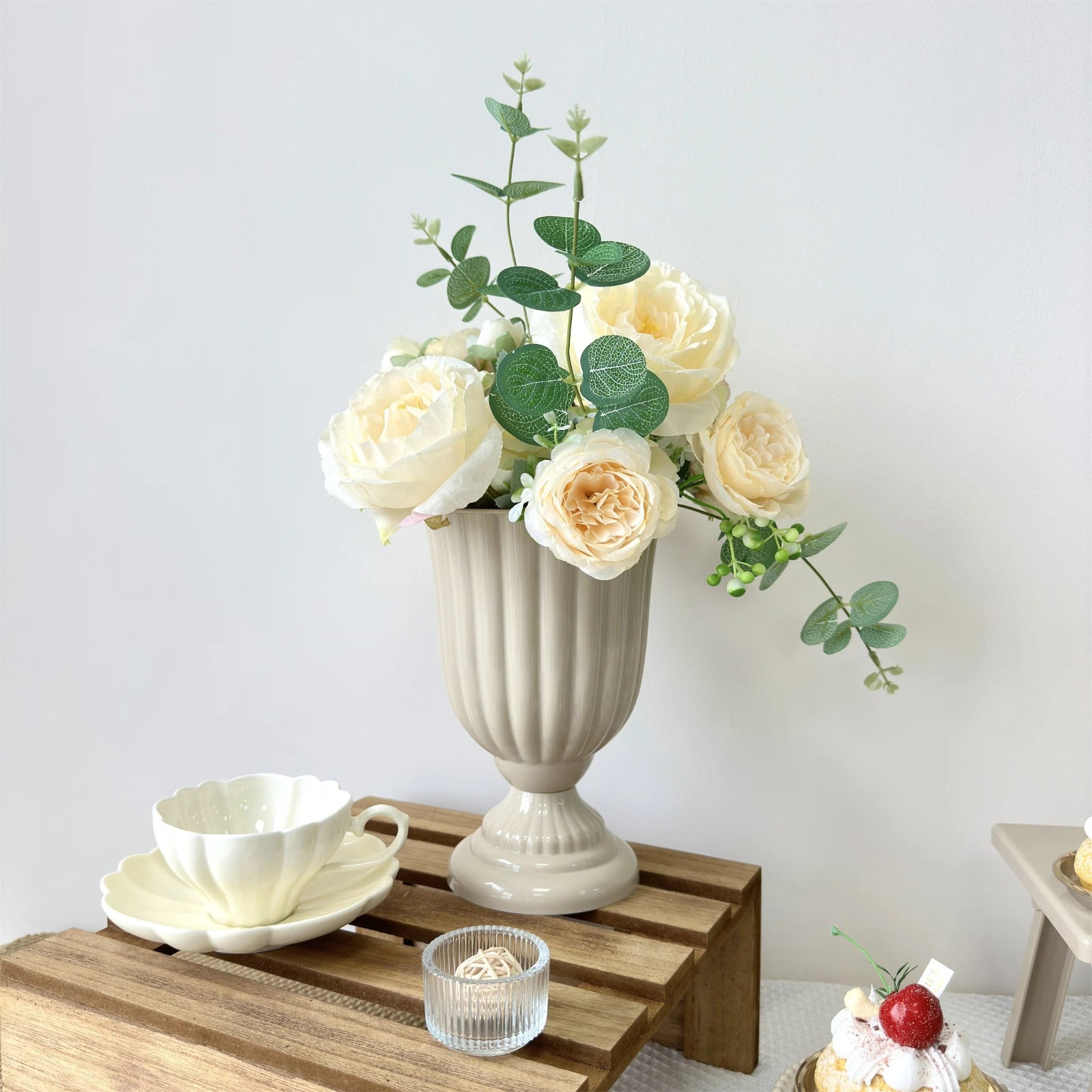 Decorative setting with a vase of flowers, teacup, and small cake on a wooden table.