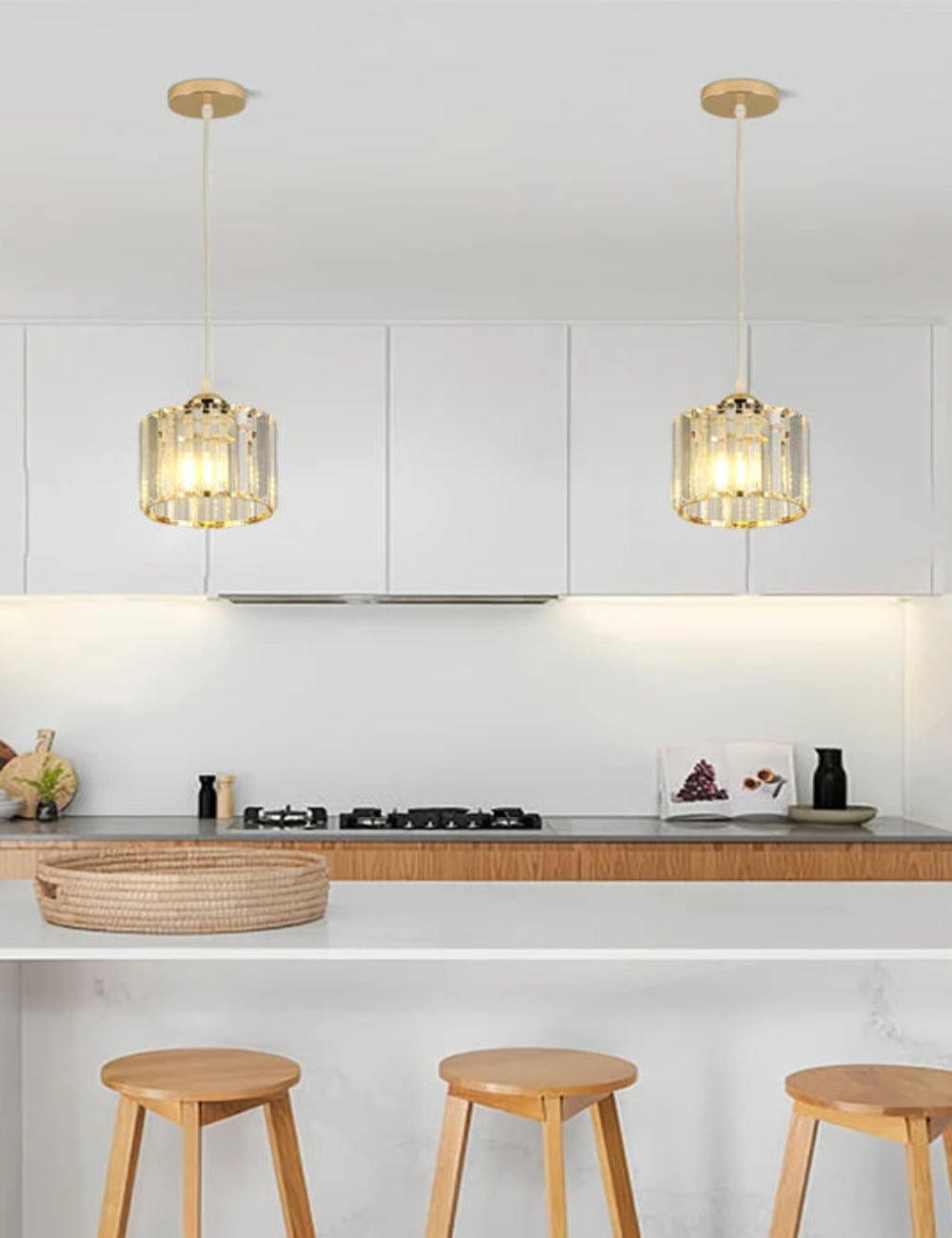 Two modern pendant lights with gold-toned metal frames and clear glass cylindrical shades hang over a white kitchen island in a bright, contemporary kitchen with light wood stools.