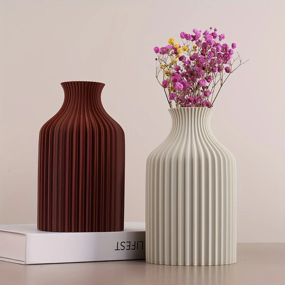 Two ribbed ceramic vases with a flared neck, one in dark reddish-brown and one in off-white, displayed on a tabletop with a white book and pink dried flowers.