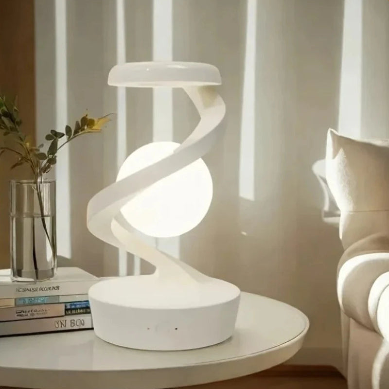 A modern white spiral-shaped table lamp with a glowing spherical bulb, placed on a round white side table in a softly lit room. A glass vase with stems and stacked books sit to the left, and part of a cream sofa is visible on the right.