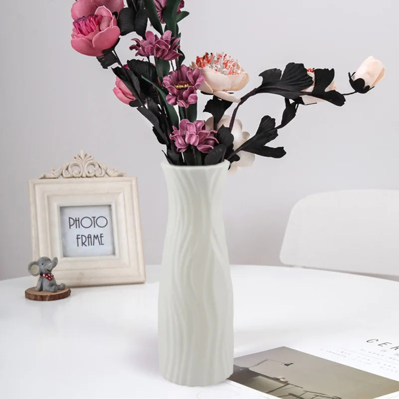 White textured vase with pink and black flowers on a white surface with a photo frame and small statue in the background.