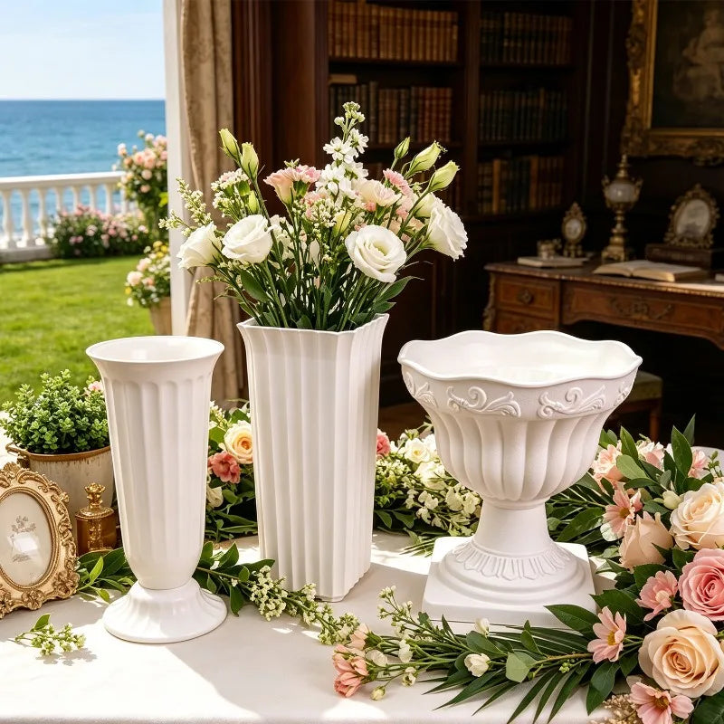 Three white decorative classical-style flower vases arranged on a table in an elegant indoor setting with a view of the ocean outside. The set includes a fluted pedestal vase, a ribbed rectangular vase with a bouquet of white and pink flowers, and a wide scalloped bowl-shaped vase surrounded by additional flowers and greenery.