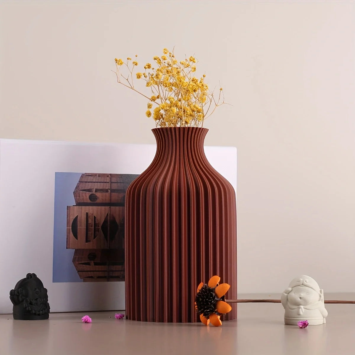 A ribbed terracotta brown ceramic vase with a flared neck, holding yellow baby's breath flowers, styled on a tabletop with a white frame, a small white figurine, and a black stone.