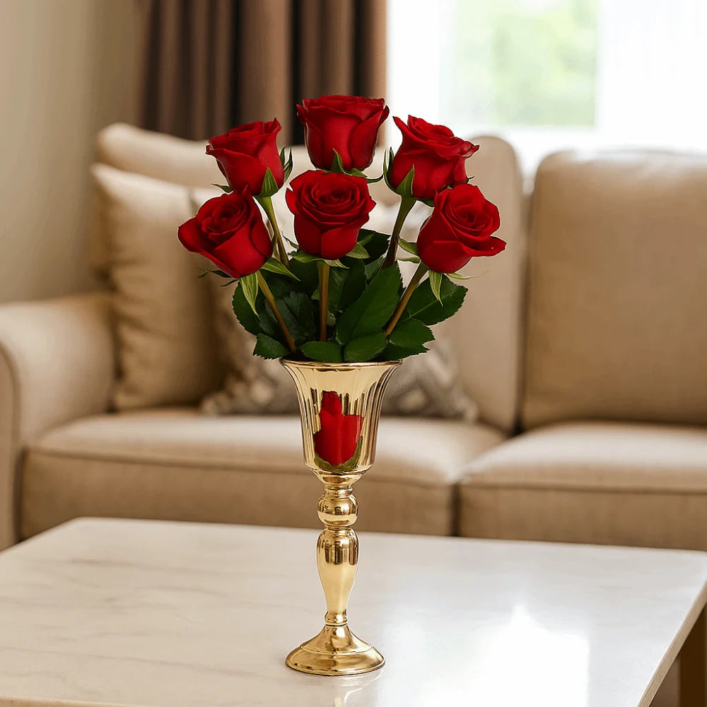 A bouquet of seven red roses with green leaves arranged in a shiny gold pedestal vase, placed on a white marble table in front of a beige sofa.