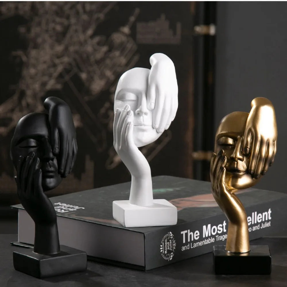 Three artistic hand and face sculptures in black, white, and gold on top of books.