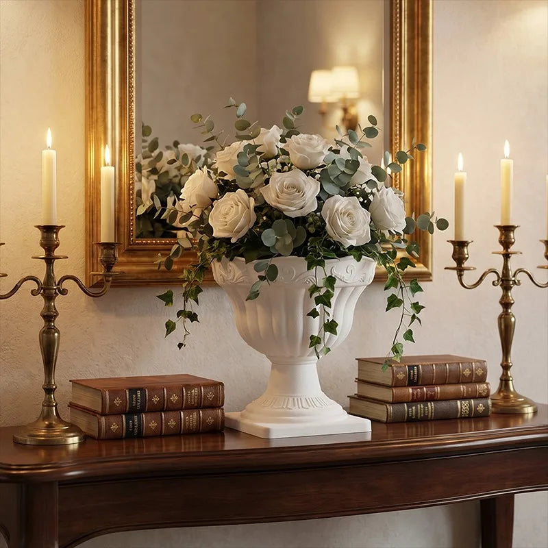 A white classical-style urn vase filled with white roses and green eucalyptus stems, displayed on a dark wooden console table. The setting is decorated with two brass candlesticks, a stack of vintage books on each side, and a large gold-framed mirror in the background.